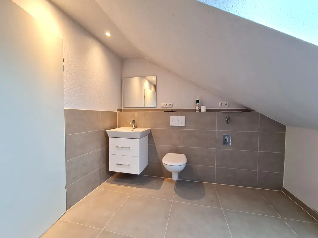 Bright bathroom with beige tile, white toilet and vanity. The ceiling slopes down on the right side.