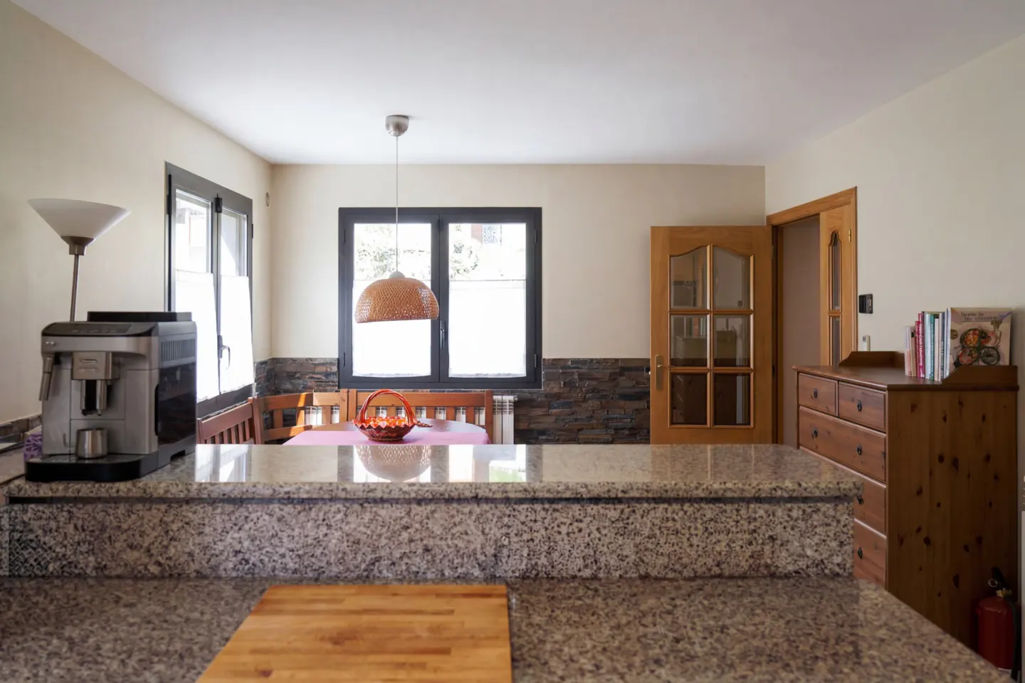 A bright kitchen with granite countertops, a coffee machine, and a dining area with a woven pendant light. A wooden door and dresser are visible.