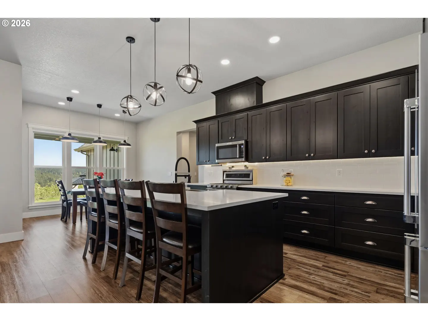 A modern kitchen with dark cabinets, a white countertop island with chairs, and wood floors.