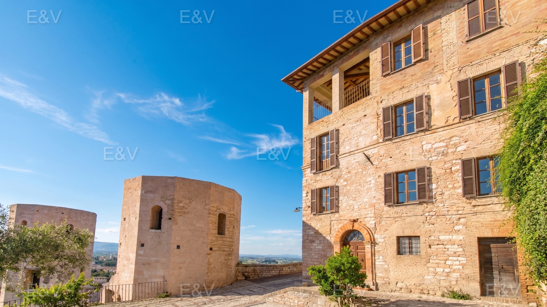 A historic stone building with brown shutters stands beside ancient stone towers under a clear blue sky.