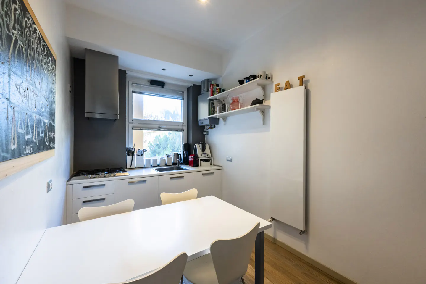 Bright kitchen with white cabinets, stainless steel hood, and a white table with chairs. A chalkboard hangs on the wall.