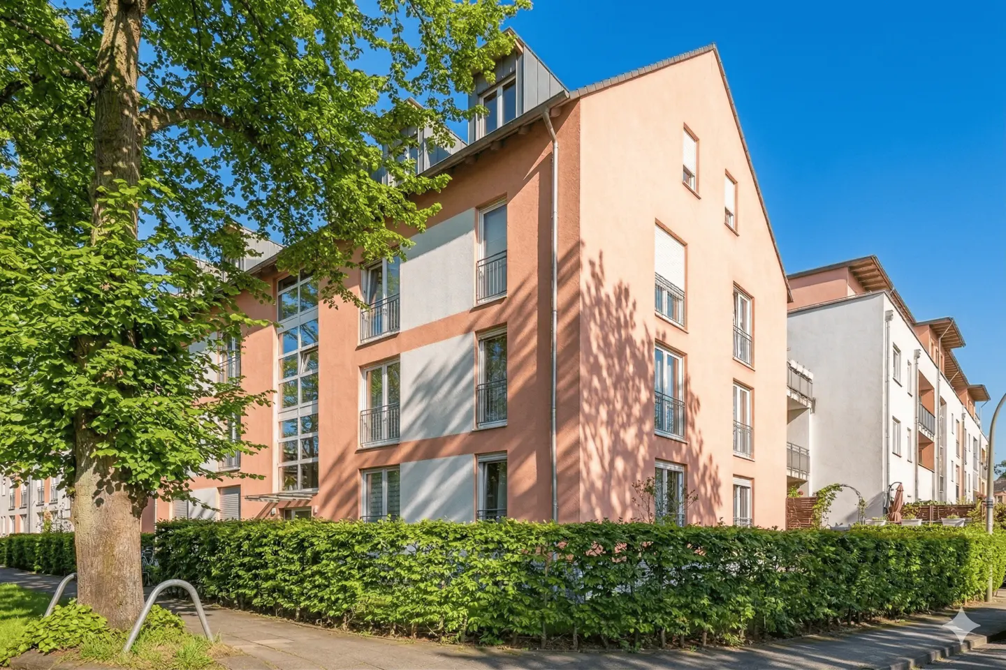 Exterior view of a peach-colored apartment building with white trim, balconies, and a green hedge in front.