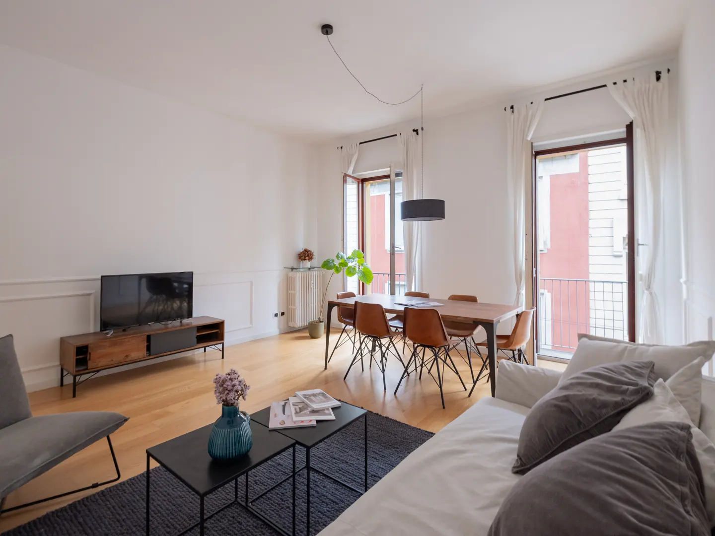 Bright living room with wood floors, white walls, and modern furniture. A dining table sits near open balcony doors with sheer curtains. A sofa and TV are in the foreground.