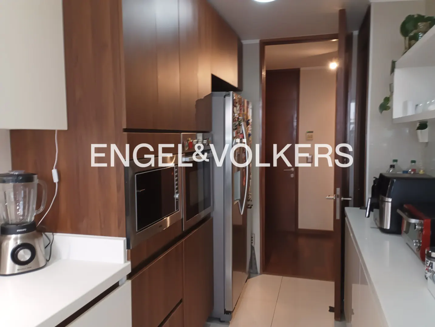A modern kitchen with wood cabinets, stainless steel appliances, and white countertops. A doorway leads to another room.