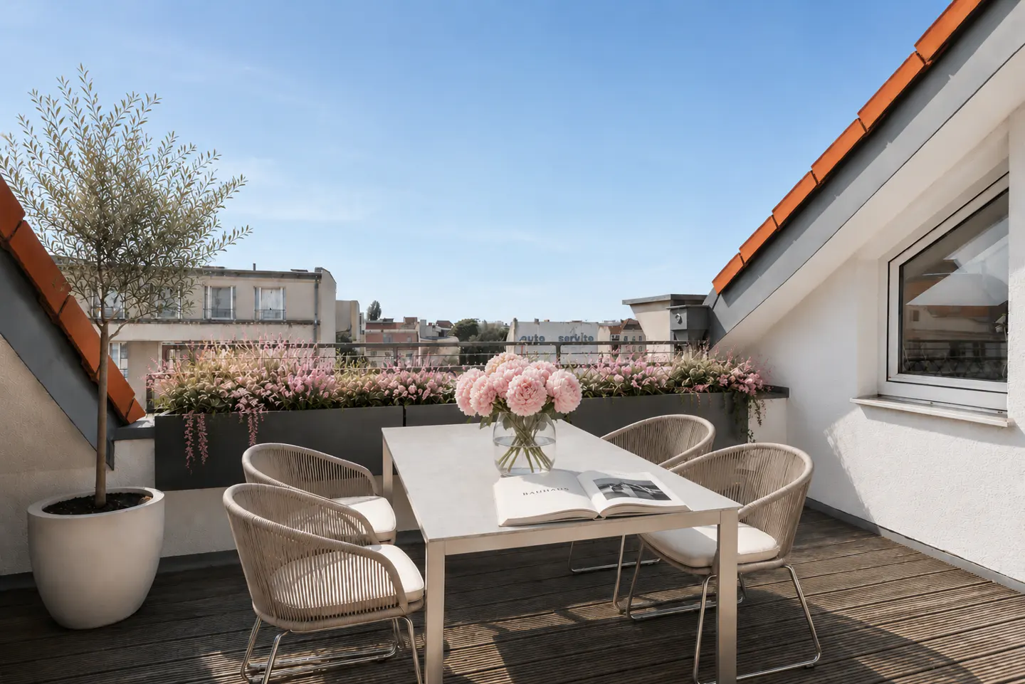 Rooftop patio with table, chairs, and flowers. A book and vase of pink flowers sit on the table. City buildings in the background.