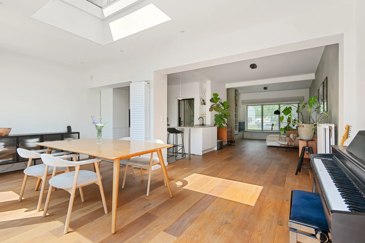 Bright open-plan living area with a wooden dining table, white chairs, a piano, and a modern kitchen in the background.
