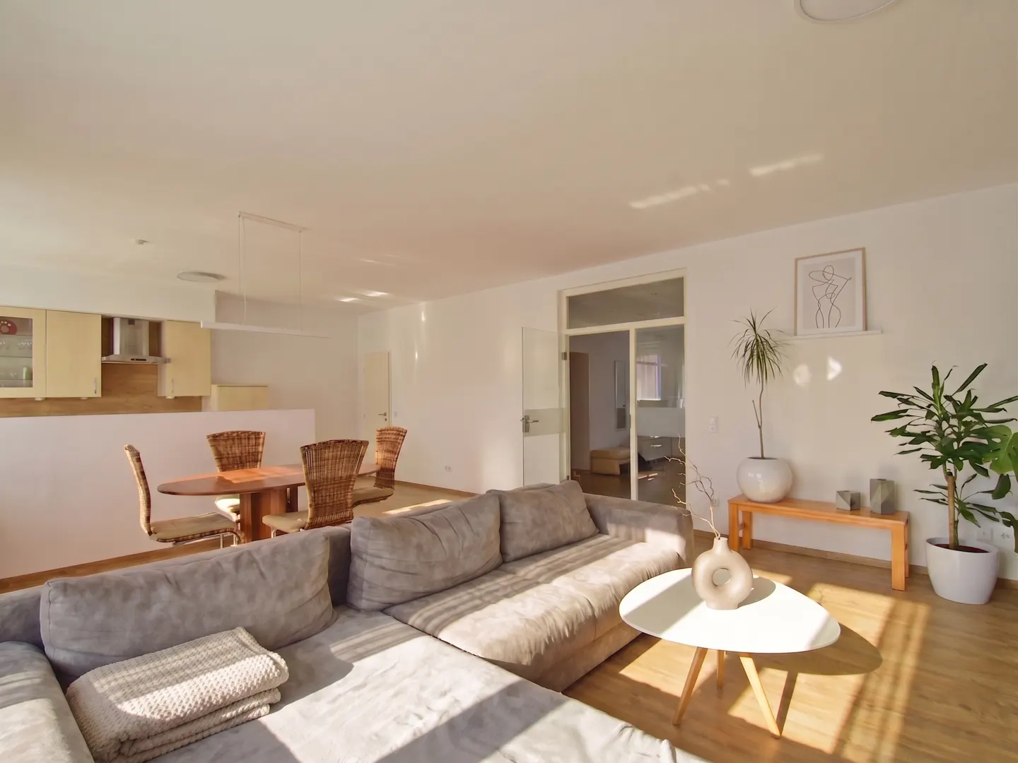 Bright, open-concept living space with a gray sectional sofa, round table, and dining area with wicker chairs. Plants add a touch of nature.