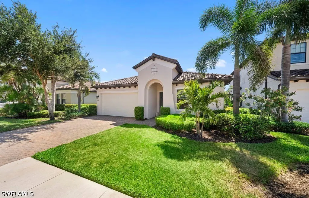 Single-story beige stucco home with a tile roof, paved driveway, manicured lawn, and palm trees under a clear blue sky.