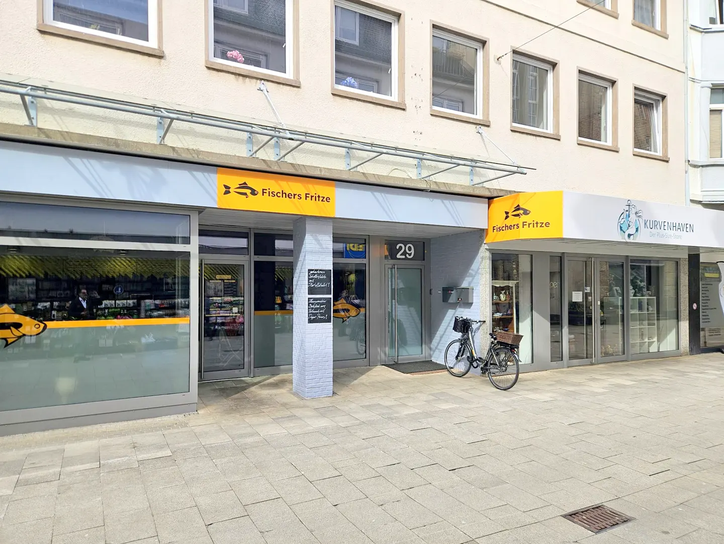 Street view of "Fischers Fritze" and "Kurvenhaven" shops with glass windows and doors, a bicycle parked outside.
