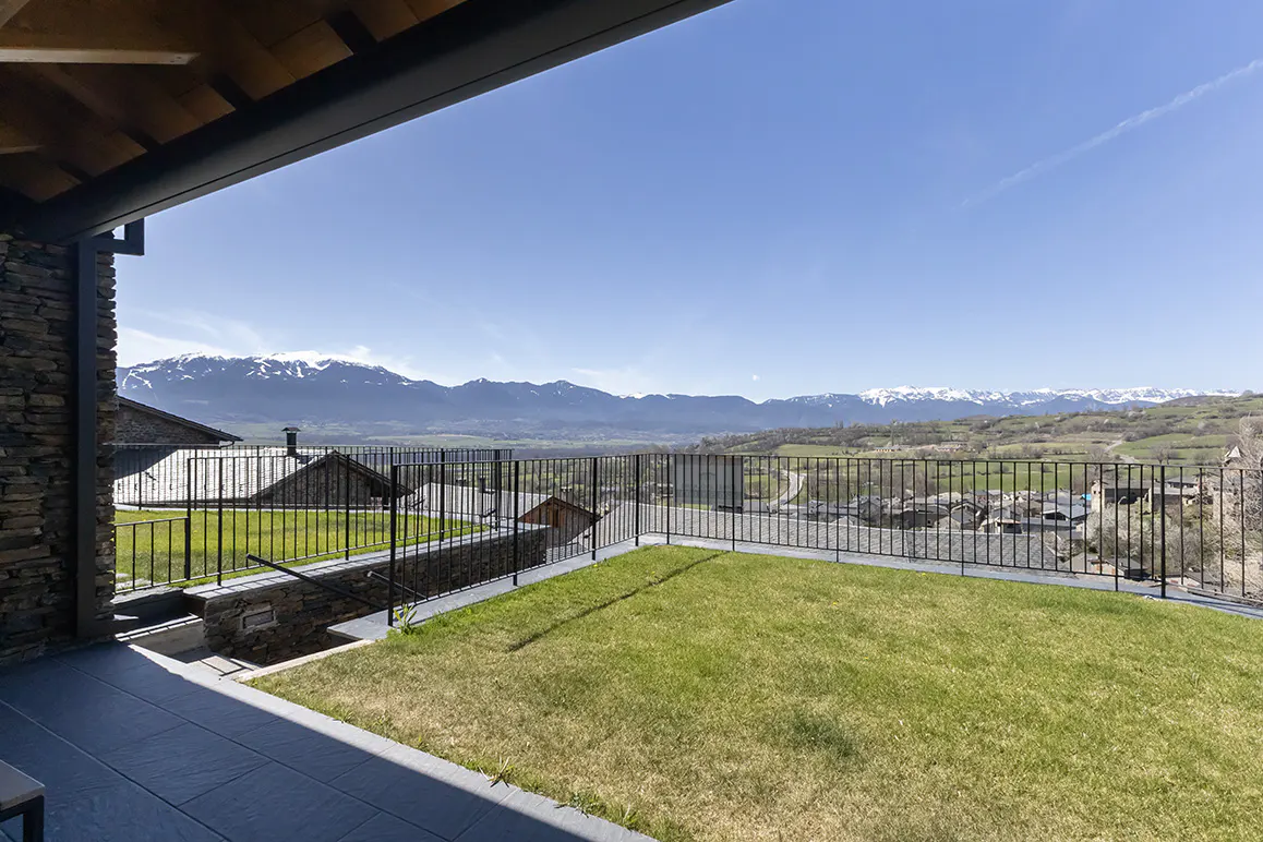 View from a stone patio with green grass, black railing, and snow-capped mountains in the background under a clear blue sky.