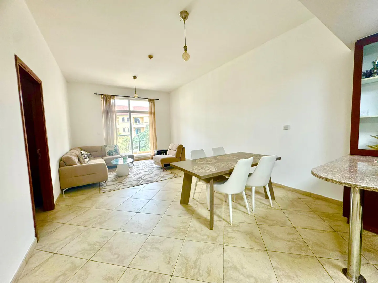 Bright living and dining area with beige sectional sofa, wood table with white chairs, and tiled floor.