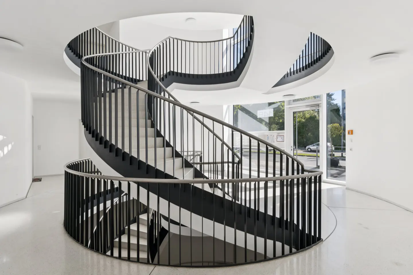 A modern, white interior features a spiral staircase with black railings and white steps. A glass door is visible in the background.