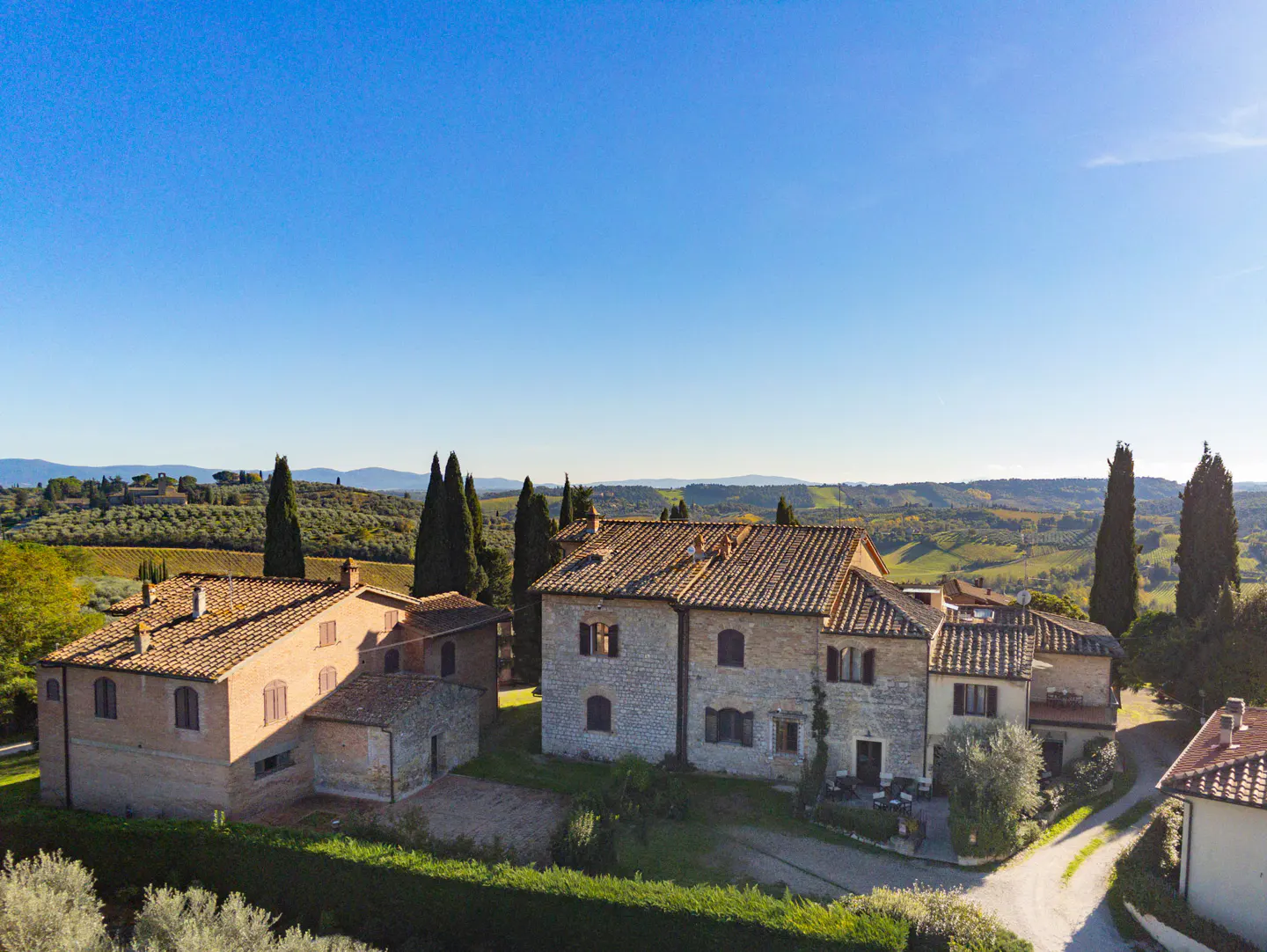 Tuscan villa with stone walls and terracotta roof tiles, surrounded by green hills and cypress trees under a clear blue sky.