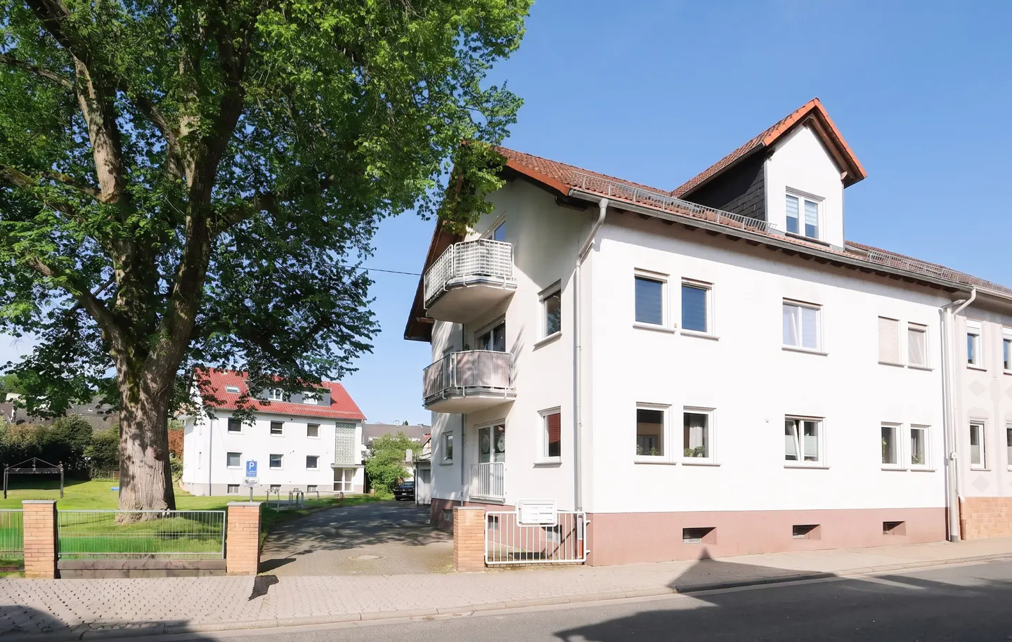Exterior view of a white apartment building with balconies and a red-tiled roof, under a blue sky. A large tree stands to the left.
