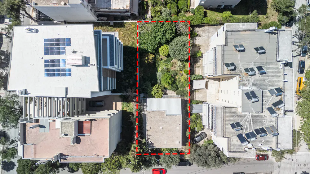Aerial view of a rectangular property outlined in red, surrounded by buildings and greenery. Solar panels are visible on rooftops.