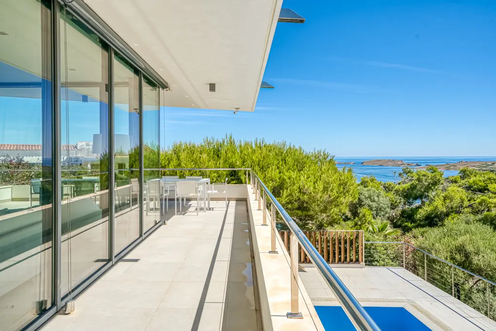 Modern balcony with glass doors, stainless steel railing, and ocean view. White table and chairs are visible.