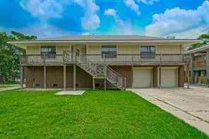 Two-story beige house with a wooden deck, stairs, and two garage doors. Green lawn and blue sky with clouds.