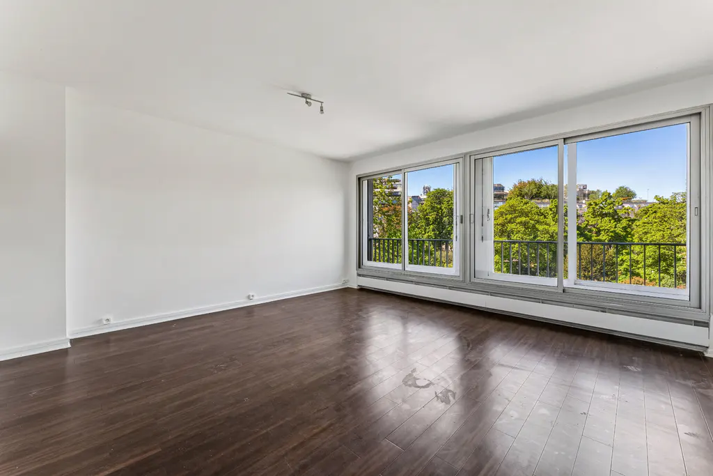 Bright, empty room with dark wood floors, white walls, and large windows overlooking trees and a blue sky.
