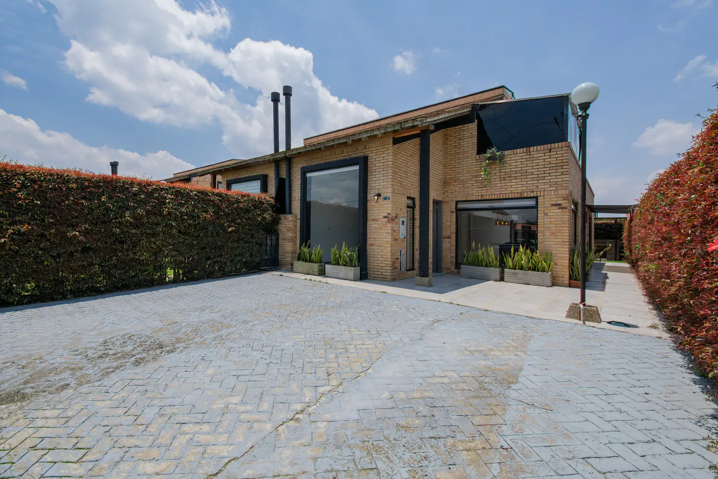 Brick house with black trim, large windows, and a gray brick driveway. Hedges line the property under a blue sky with clouds.
