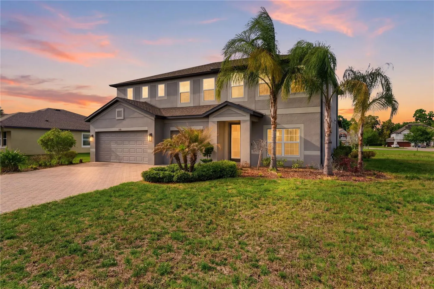 Two-story gray house with a two-car garage, palm trees, and a green lawn at sunset.
