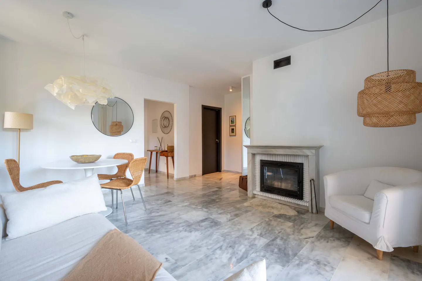 Bright living room with marble floors, white walls, fireplace, white sofa, round table with wicker chairs, and wicker pendant light.