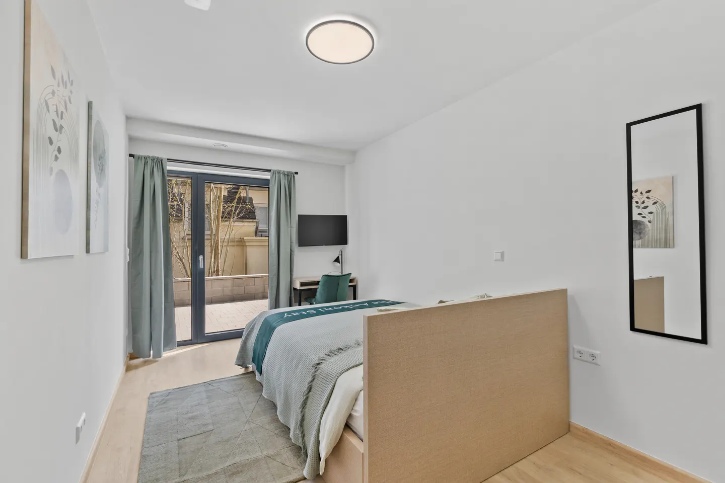 Bright bedroom with white walls, light wood floors, and a bed with a blue and gray blanket. A desk, TV, and patio doors are visible.
