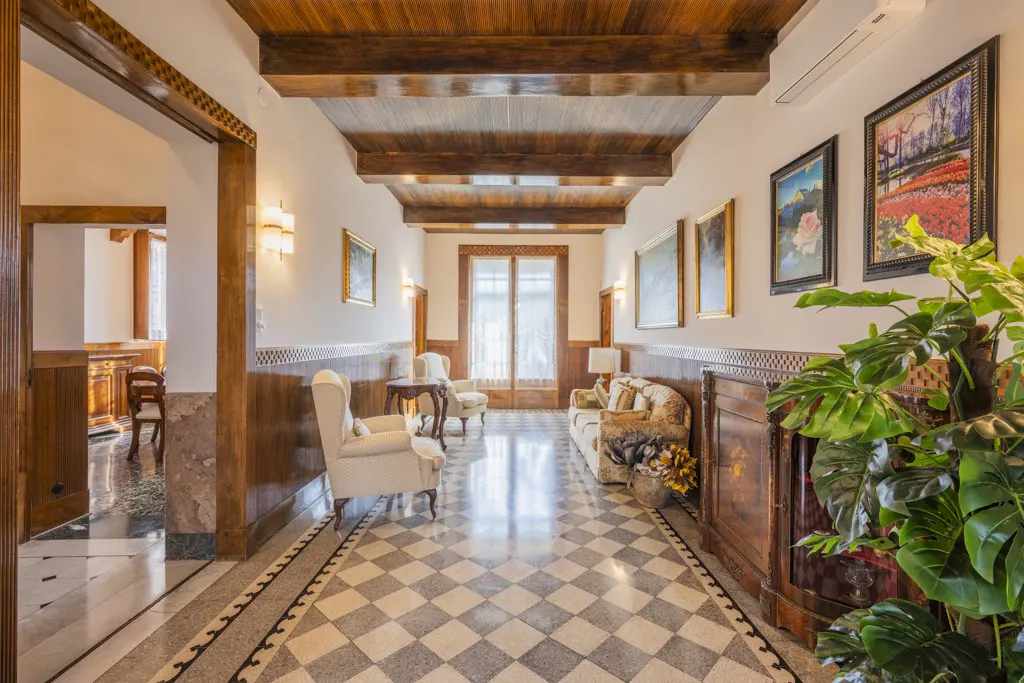 Hallway with checkered floor, wood paneled walls, and beamed ceiling. Furniture includes chairs, sofa, and paintings on the wall.