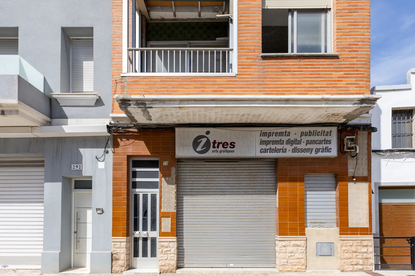 A two-story building with a brick facade and a closed metal shutter. A sign reads "Z tres arts grafiques". A small balcony is visible on the second floor.