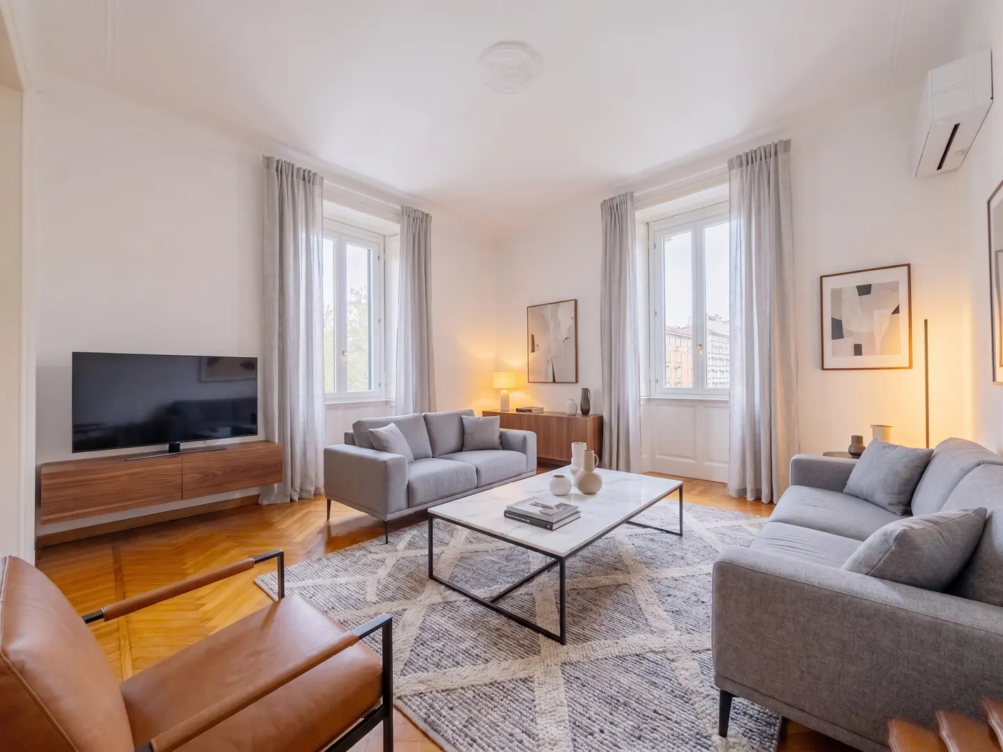 Bright living room with two gray sofas, a marble coffee table, and a brown leather chair on a patterned rug. Two windows with gray curtains.