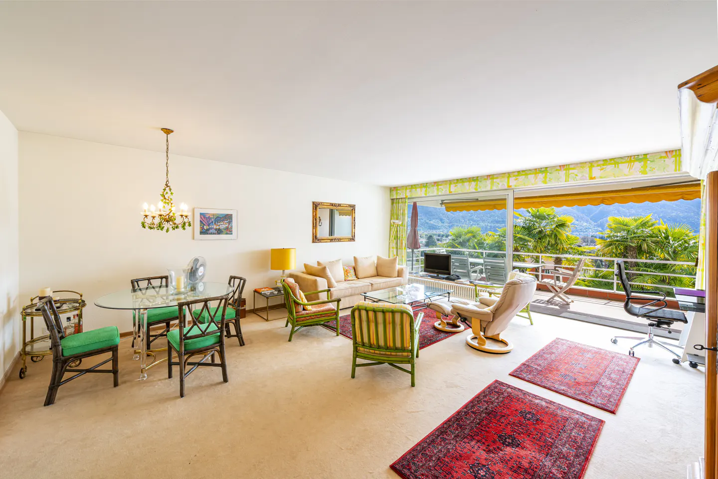 Bright living room with a glass table, green chairs, a beige sofa, and a balcony with a mountain view.
