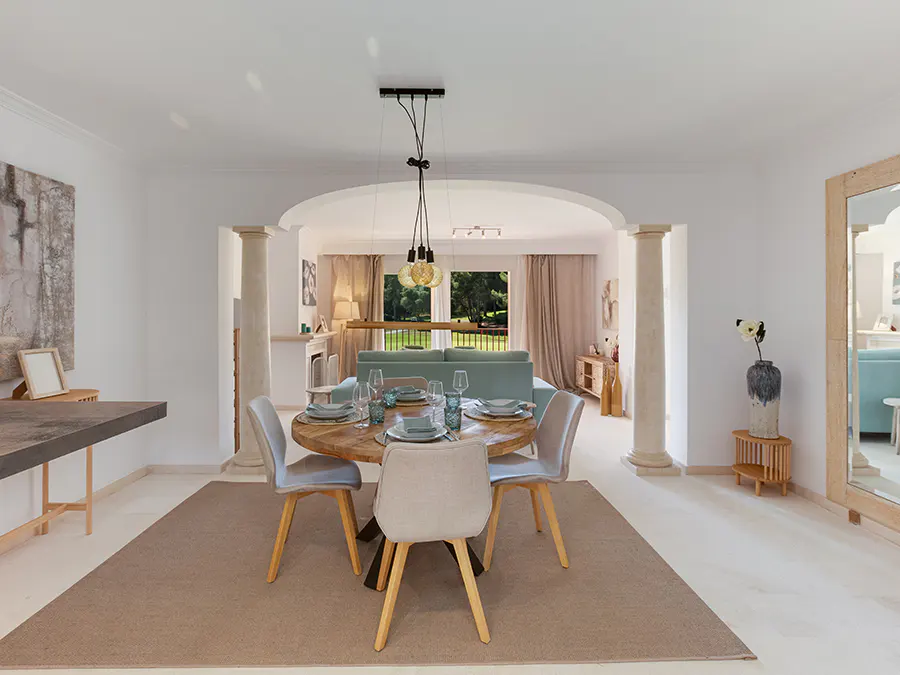 Bright, open dining room with a round wooden table set for four. Light gray chairs, tan rug, and white walls. Living room visible beyond archway.