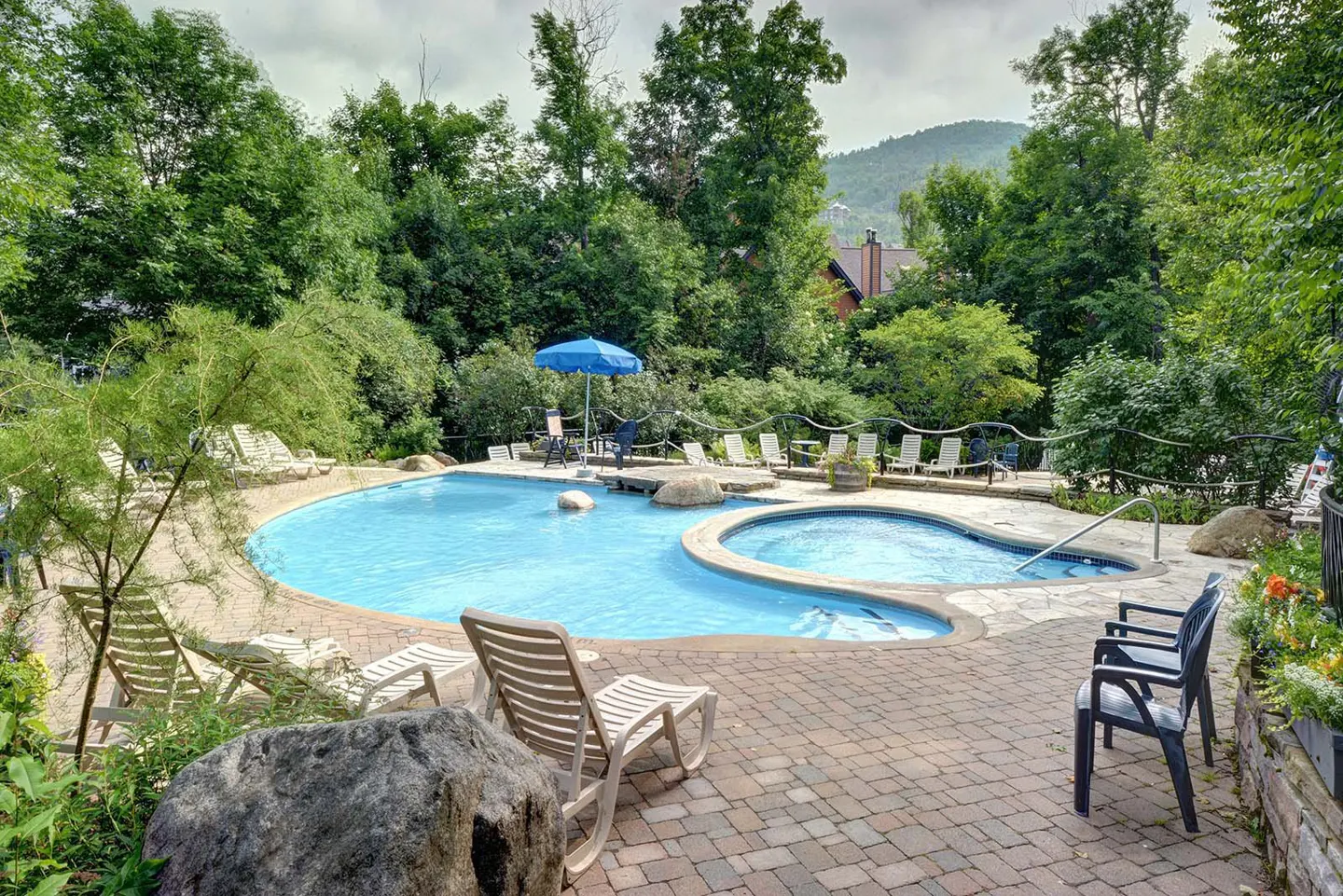Outdoor pool area with lounge chairs, a blue umbrella, and a hot tub, surrounded by lush green trees and a brick patio.