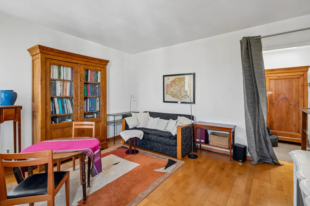 Living room with a dark sofa, wooden bookcase, dining table with a pink cloth, and an orange patterned rug on wood floors.