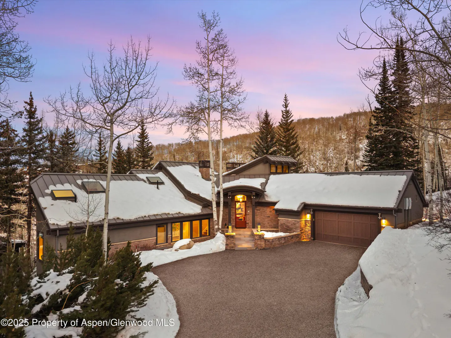 Exterior view of a luxury home with snow-covered roof and driveway at dusk. Trees surround the house.