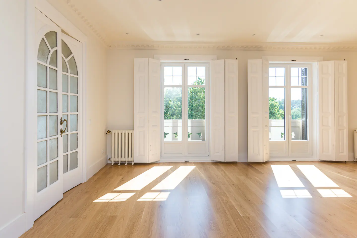 Bright, empty room with light wood floors, white walls, arched glass doors, and two windows with open white shutters.