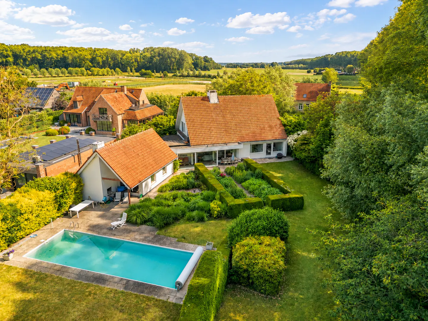 Aerial view of a house with a red-tiled roof, a swimming pool, and a lush garden, set against a scenic rural landscape.