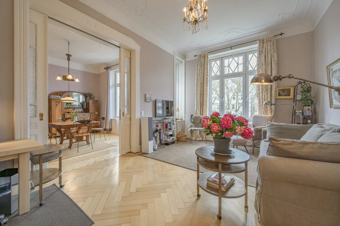 A bright living room with a herringbone wood floor, a sofa, a round table with pink flowers, and a view into the dining room.