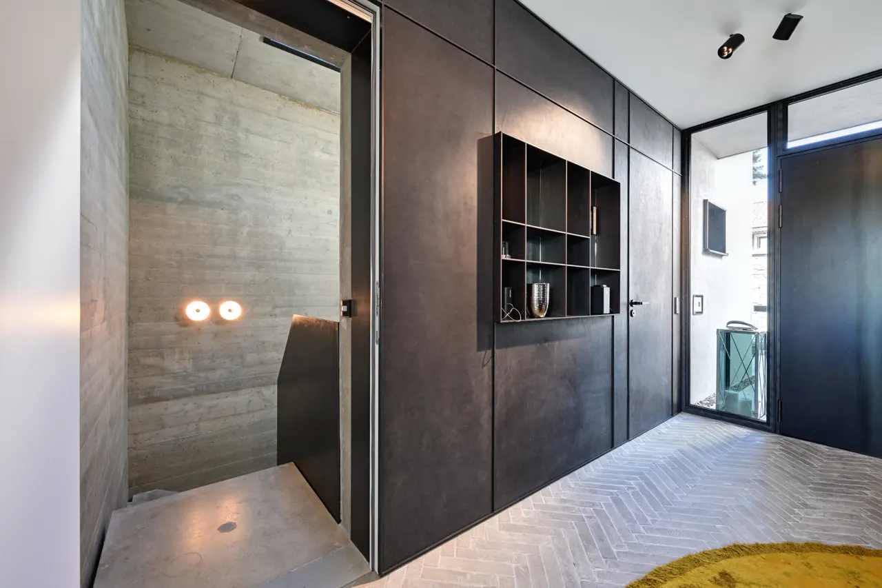Modern hallway with herringbone floors, black paneled walls, and a concrete staircase. A black shelf displays decorative objects.