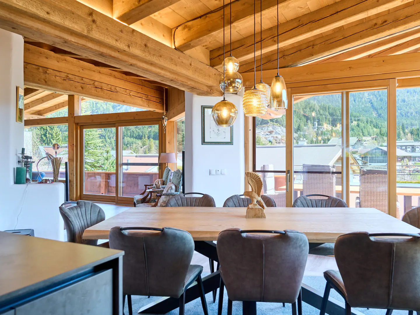 A dining room with a wood table, gray chairs, and pendant lights. Large windows offer a view of mountains and trees.