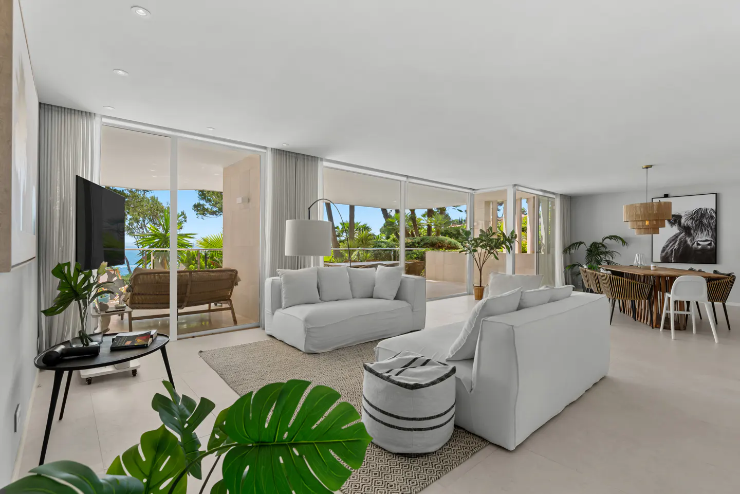 Bright, open-plan living room with white sofas, dining table, and outdoor views. A modern, airy space with neutral tones and natural light.