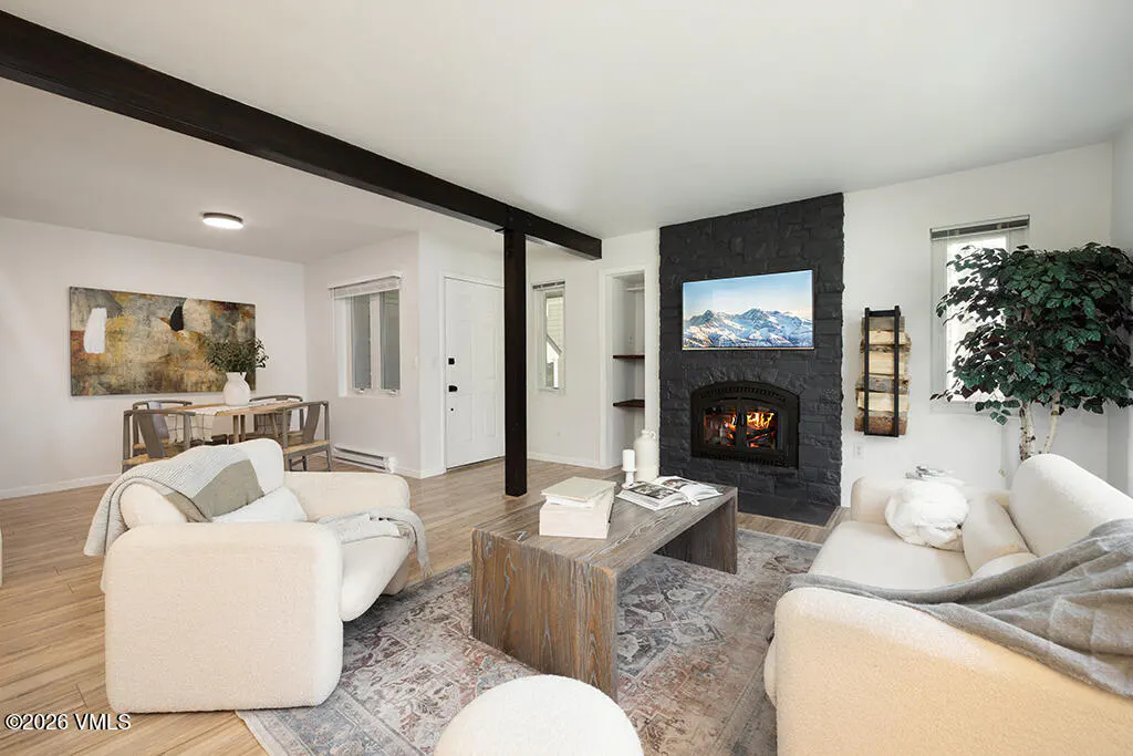Living room with white walls, wood floors, and black fireplace. A TV hangs above the fireplace, showing a mountain scene. Two white chairs and a wood coffee table sit on a patterned rug.