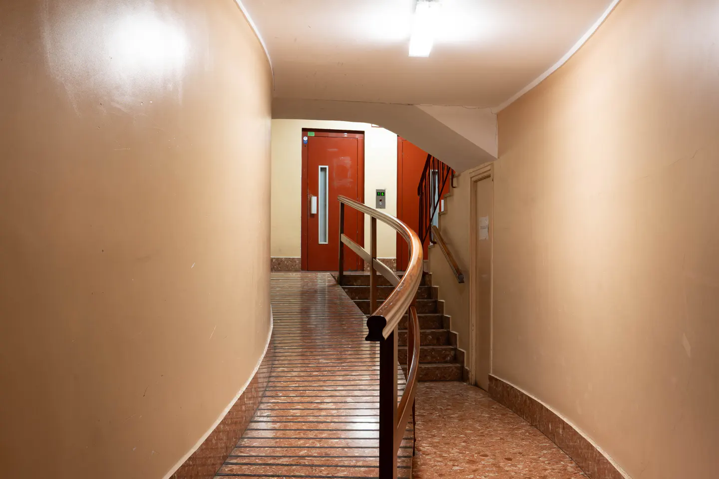 Hallway with beige walls, a red elevator door, and stairs with a wooden handrail. The floor is tiled in red and brown.