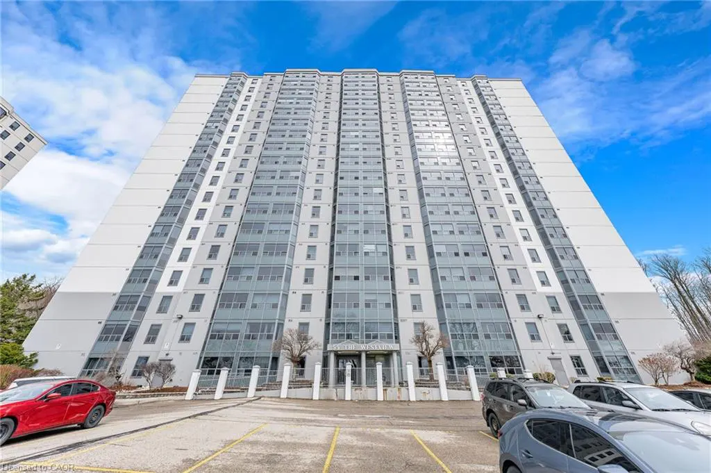 Exterior view of a tall, gray apartment building with many windows, a parking lot, and cars under a blue sky.
