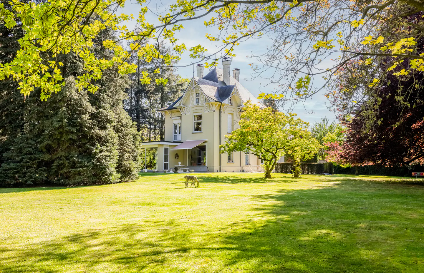 A pale yellow Victorian house with a large green lawn and trees. A deer grazes in the yard.