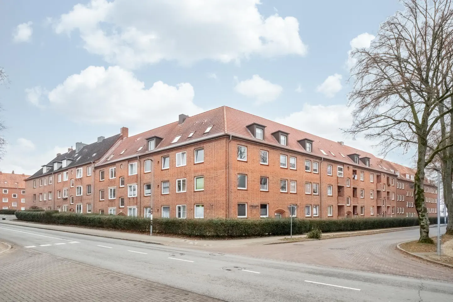 Exterior view of a long, red brick apartment building with a red tile roof and white-framed windows. A green hedge runs along the base.