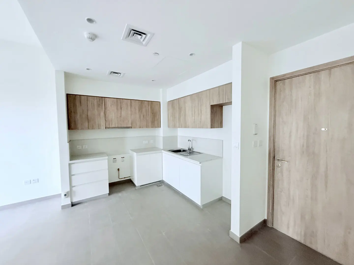 A bright, empty kitchen with white cabinets, wood-look upper cabinets, gray floors, and a closed wood-look door.