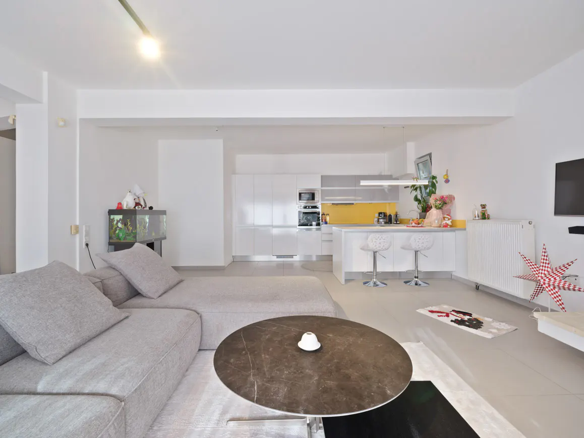 Modern open-plan living room with a grey sectional sofa, round marble coffee table, and kitchen with yellow backsplash.