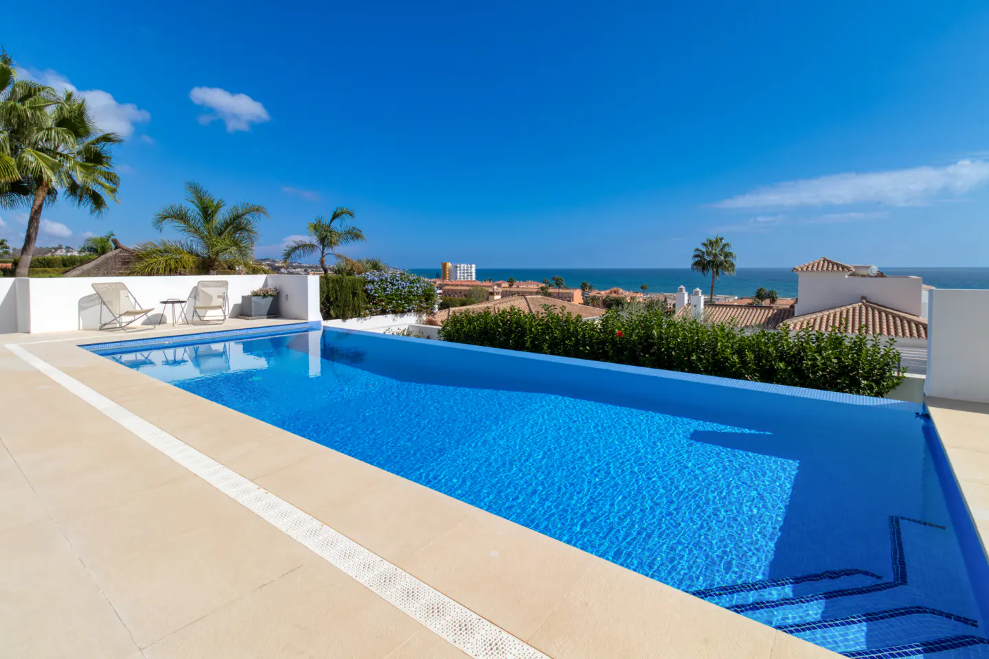 Infinity pool with blue tiles overlooking the ocean. Palm trees and buildings in the background under a clear blue sky.