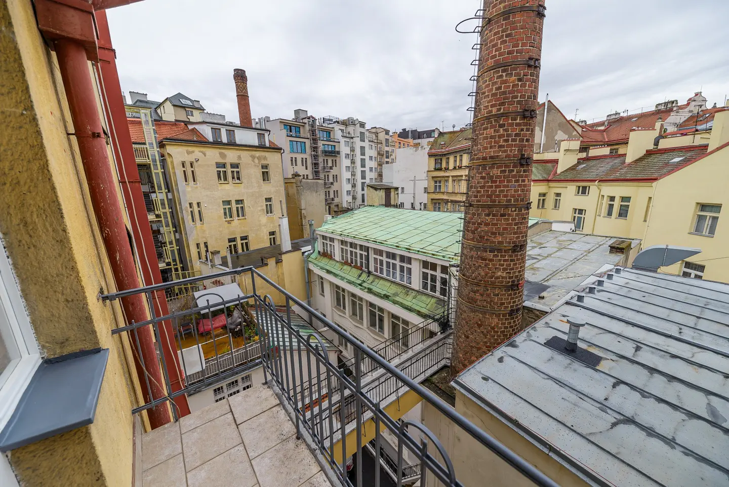View from a balcony overlooking a European city with yellow buildings, a brick chimney, and a cloudy sky.