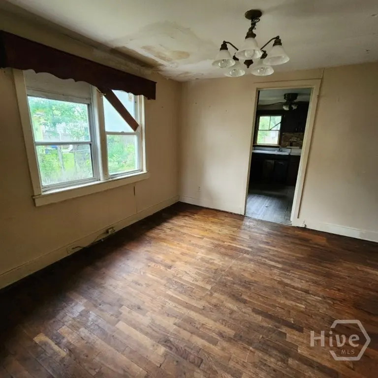 A room with hardwood floors, a window with a broken valance, and a doorway to a kitchen.